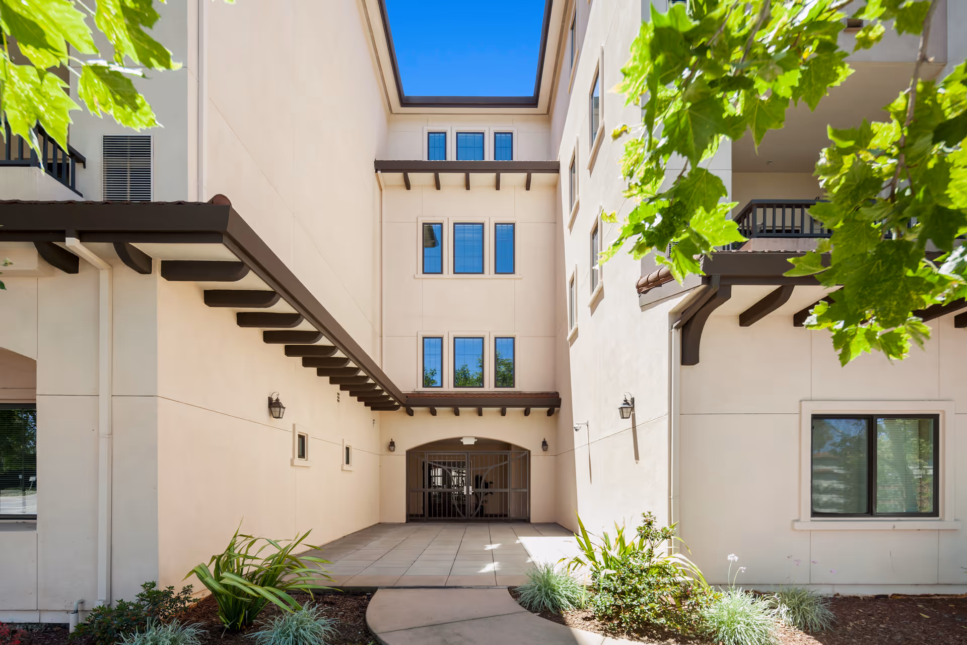 Entrance courtyard of a beige stucco building with a gated passage, balconies, landscaping and a bright blue sky.