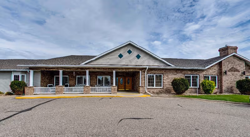 Front exterior view of a single-story brick building with a covered entrance supported by white columns, a paved driveway, and a partly cloudy sky above.