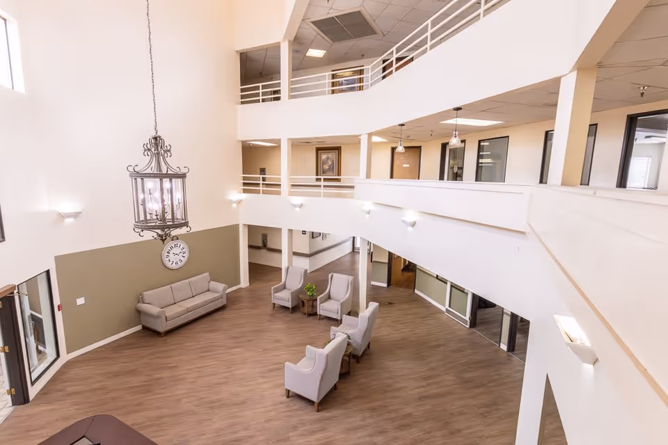 Spacious two-story common area in a senior living facility with wood flooring, beige walls, and modern lighting. The area features a beige sofa, four armchairs arranged around two small tables with a plant, a large hanging chandelier, and a wall clock. The upper level has a white railing overlooking the seating area below.