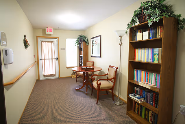 Carpeted hallway/common sitting area with a small round table and two chairs, bookshelves, a floor lamp, and an exit door.