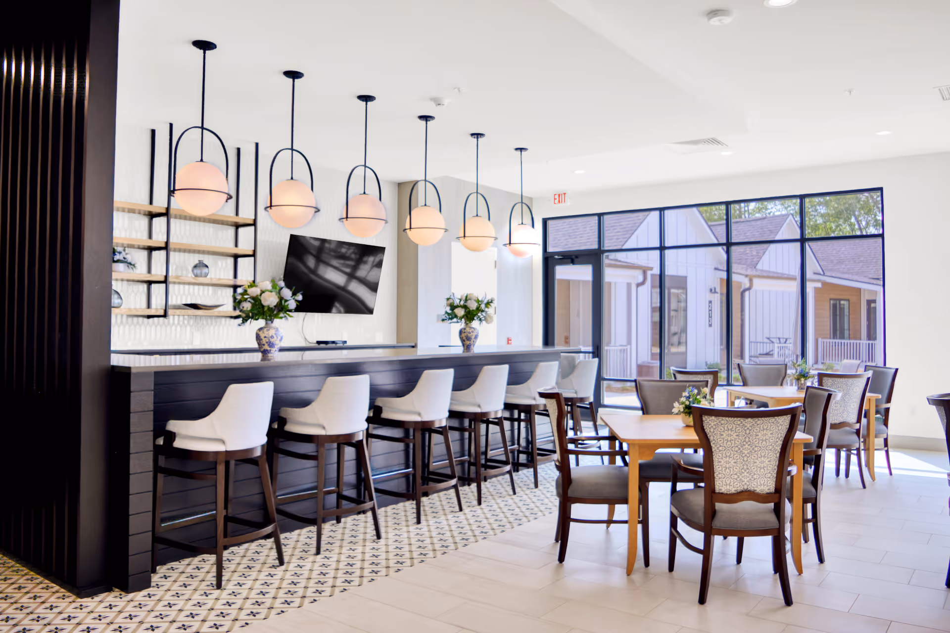 A bright and modern dining area with a long black bar counter featuring six white cushioned bar stools. Above the counter hang six round pendant lights with black frames. Behind the bar is a wall-mounted flat-screen TV and open shelves with decorative items. To the right, there are wooden dining tables with upholstered chairs, each table decorated with a small floral arrangement. Large windows in the background let in natural light and offer a view of an outdoor patio area with buildings.