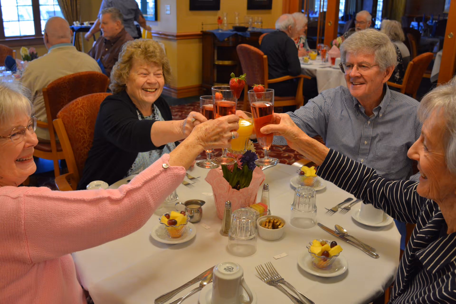 Four elderly people sitting around a dining table in a restaurant or dining room, smiling and raising glasses filled with a pink beverage garnished with strawberries in a toast. The table is set with plates of fruit, utensils, glasses, and a small flower arrangement in the center. Other diners are visible in the background.