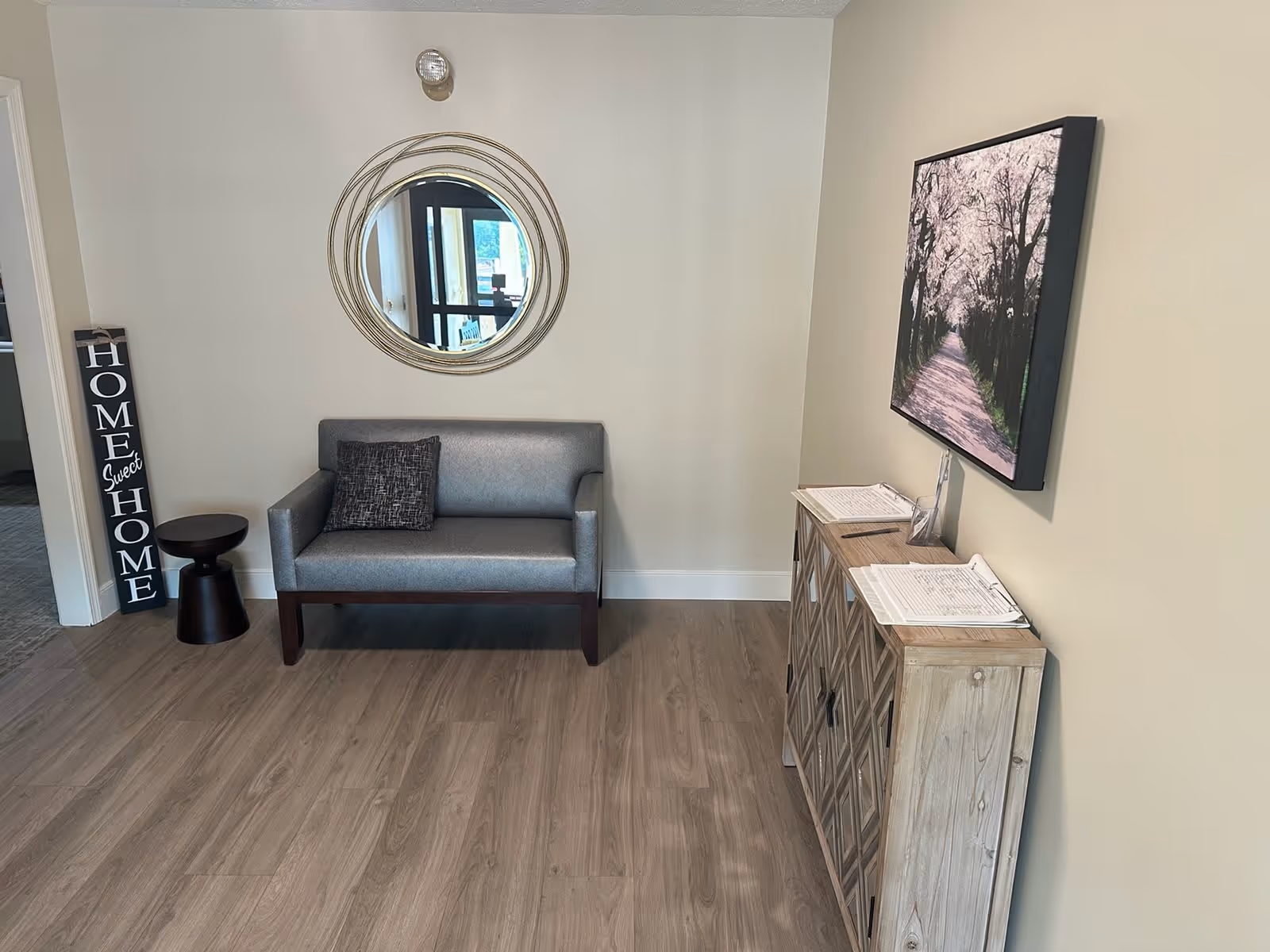 A small seating area in a room with light-colored walls and wood flooring. There is a gray loveseat with a dark pillow, a small round black side table, and a decorative sign leaning against the wall that reads 'HOME Sweet HOME'. Above the loveseat is a round mirror with a decorative frame. To the right, there is a wooden cabinet with a geometric pattern on the doors, topped with clipboards and a pen. A framed picture of a tree-lined path hangs above the cabinet.