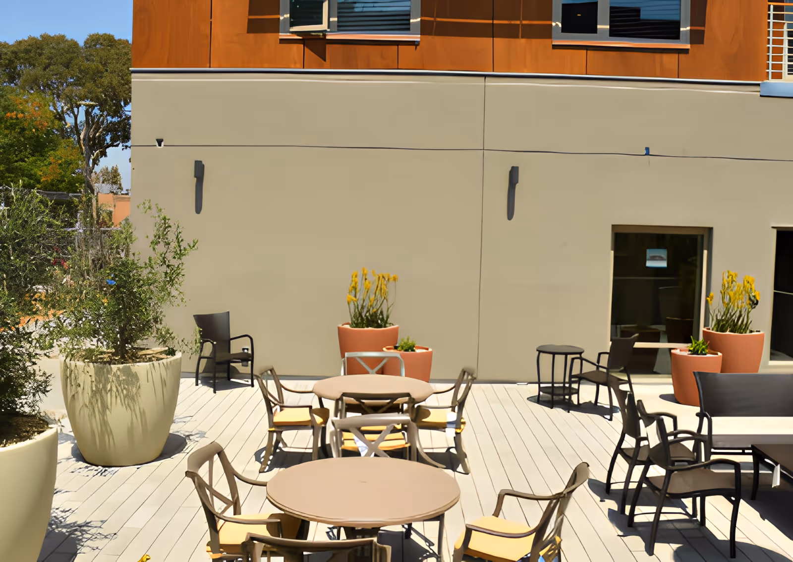 Outdoor patio area with multiple round tables and chairs arranged on a wooden deck. Large potted plants with greenery and yellow flowers are placed around the space. The background shows a beige and orange building wall with windows.