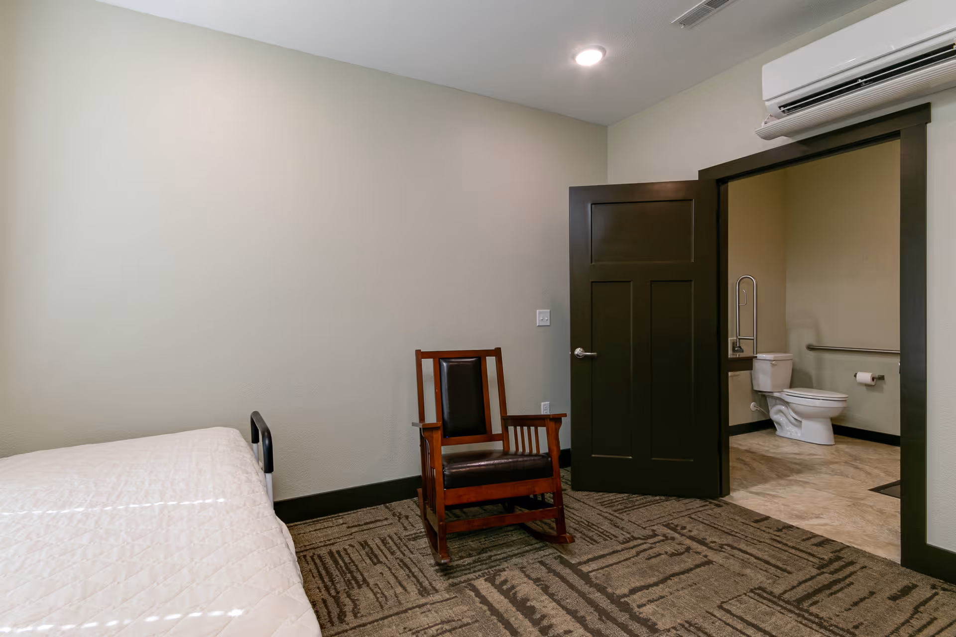 Simple bedroom with a bed, a wooden chair, and an open door revealing a bathroom with a toilet.