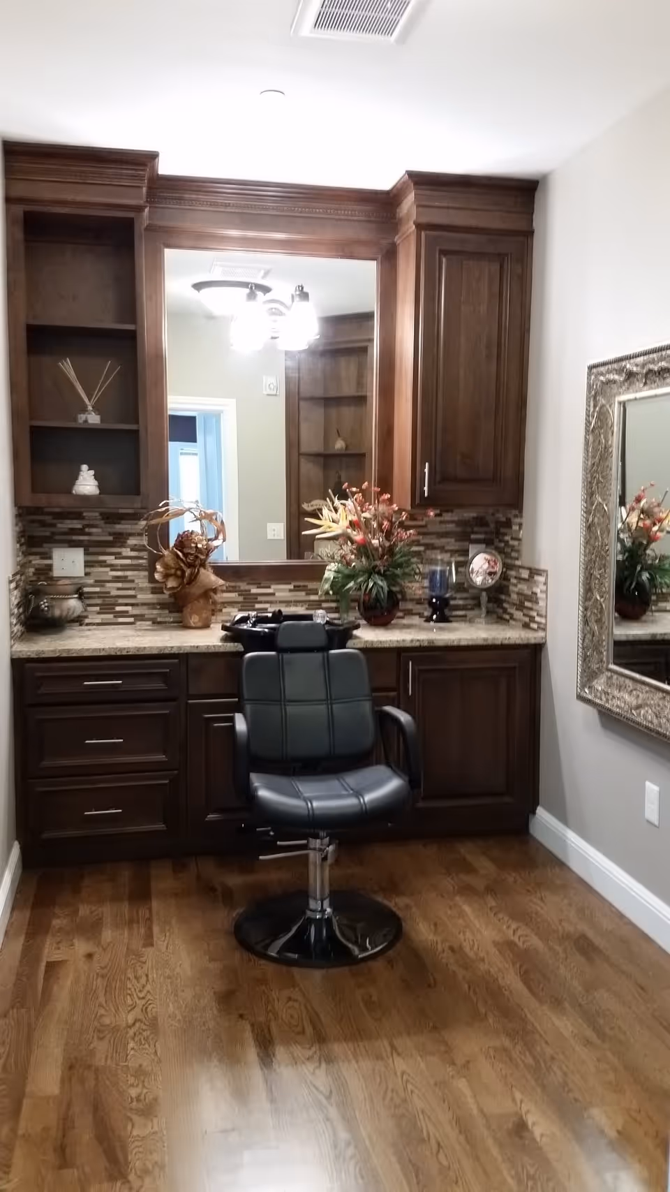A room with wooden cabinets and a countertop featuring decorative items and a large mirror. In front of the counter is a black salon chair on a wooden floor. A large ornate framed mirror is mounted on the right wall.