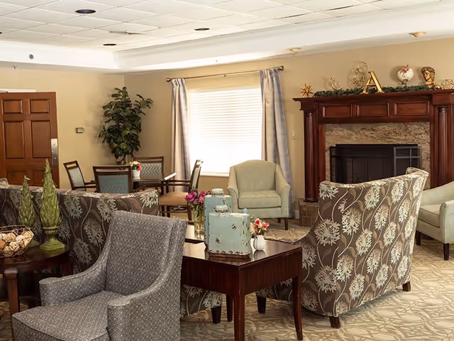 A cozy living room area with patterned armchairs, a wooden coffee table decorated with small vases of flowers, and a fireplace with a wooden mantel adorned with decorative items. There is a window with curtains letting in natural light, a potted plant in the corner, and a wooden door visible in the background.