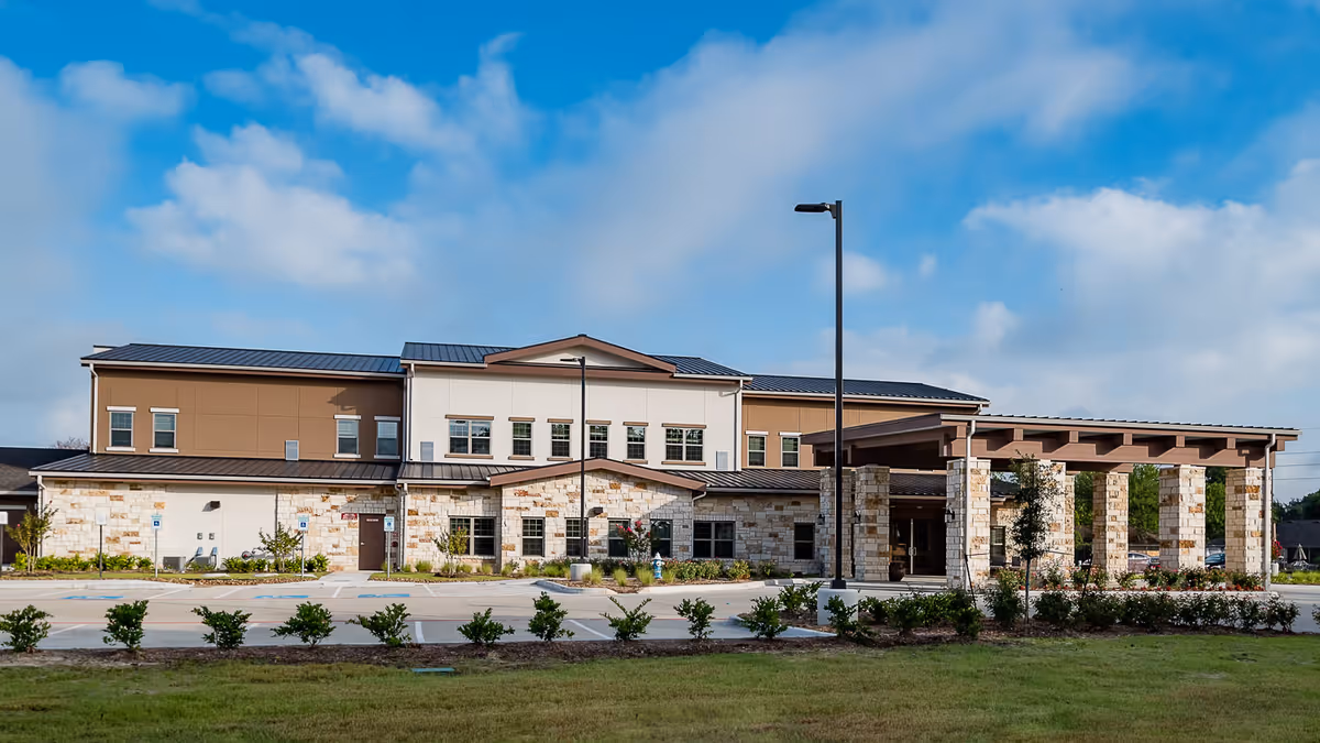 Exterior view of a modern senior living facility building with stone and beige siding, multiple windows, a covered entrance supported by stone pillars, a parking area, and landscaped greenery under a partly cloudy blue sky.