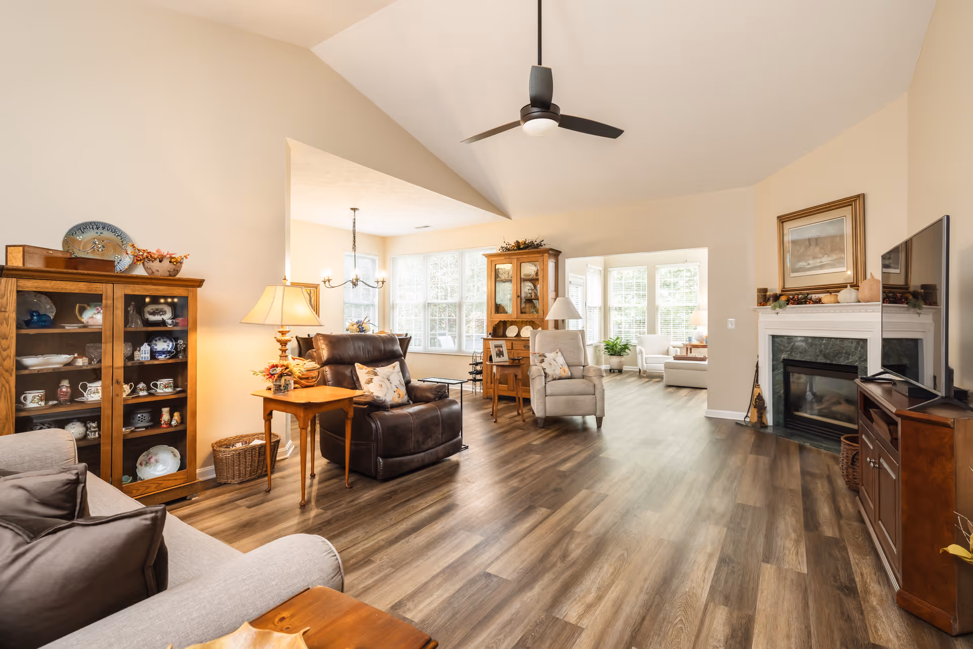 A spacious and well-lit living room area with wooden flooring, beige walls, and vaulted ceilings. The room features a variety of seating options including a gray sofa, a dark brown leather recliner, and a light gray armchair. There is a wooden display cabinet with decorative dishes, a wooden side table with a lamp, and a fireplace with a TV mounted above it. Large windows allow natural light to fill the space, and there are decorative items on the mantel and furniture.