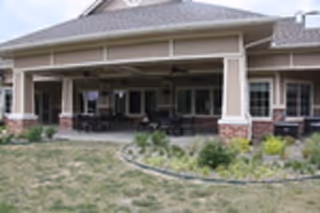 Covered outdoor patio area attached to a building with multiple windows and columns, surrounded by a garden with various plants and a grassy lawn in the foreground.
