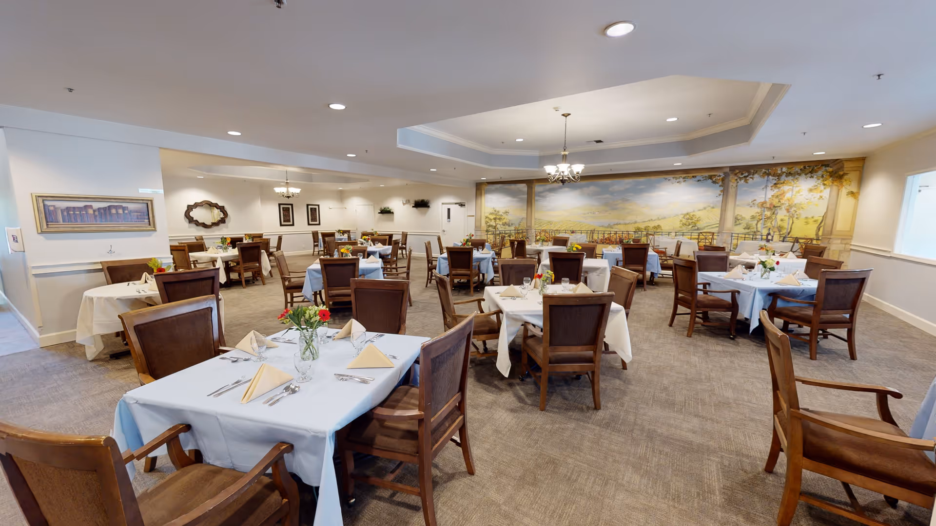 A spacious dining room with multiple tables covered in white and light blue tablecloths, each set with folded napkins, silverware, and small flower arrangements. The room features wooden chairs, carpeted flooring, and a large mural on the far wall depicting a scenic outdoor landscape. Ceiling lights and chandeliers provide illumination.