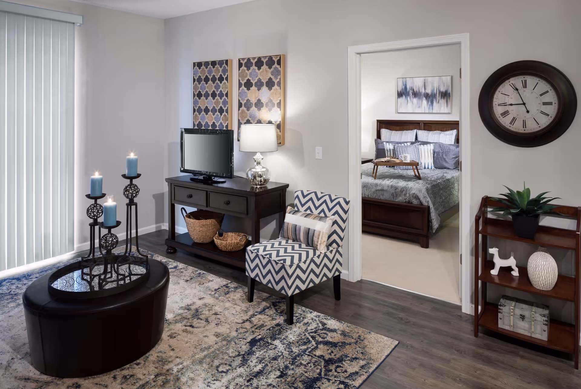 A cozy living room area with a patterned armchair, a dark wooden TV stand holding a small flat-screen TV and a lamp, and a round ottoman with a candle holder featuring four lit blue candles. The room has a large window with vertical blinds and a patterned rug on the floor. Through an open doorway, a bedroom is visible with a bed made with gray and white bedding, a wooden headboard, a small tray on the bed, and abstract artwork on the wall. A large round clock and a small wooden shelf with decorative items are on the right wall of the living room.