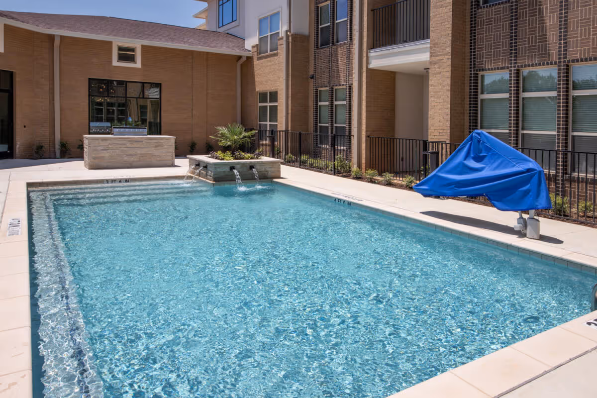 Outdoor swimming pool with clear blue water, a small water feature with three spouts, a covered pool lift with a blue cover, and a built-in grill area against a brick building under a clear sky.