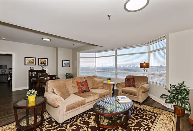 A cozy living room with a beige sofa and armchair, both adorned with decorative pillows. A glass-top coffee table sits on a patterned area rug. Large windows with blinds offer a panoramic view outside. There is a side table with a yellow pot plant, a floor lamp, and additional plants near the windows. In the background, a dining area with a table and chairs is visible, along with framed pictures on the wall.