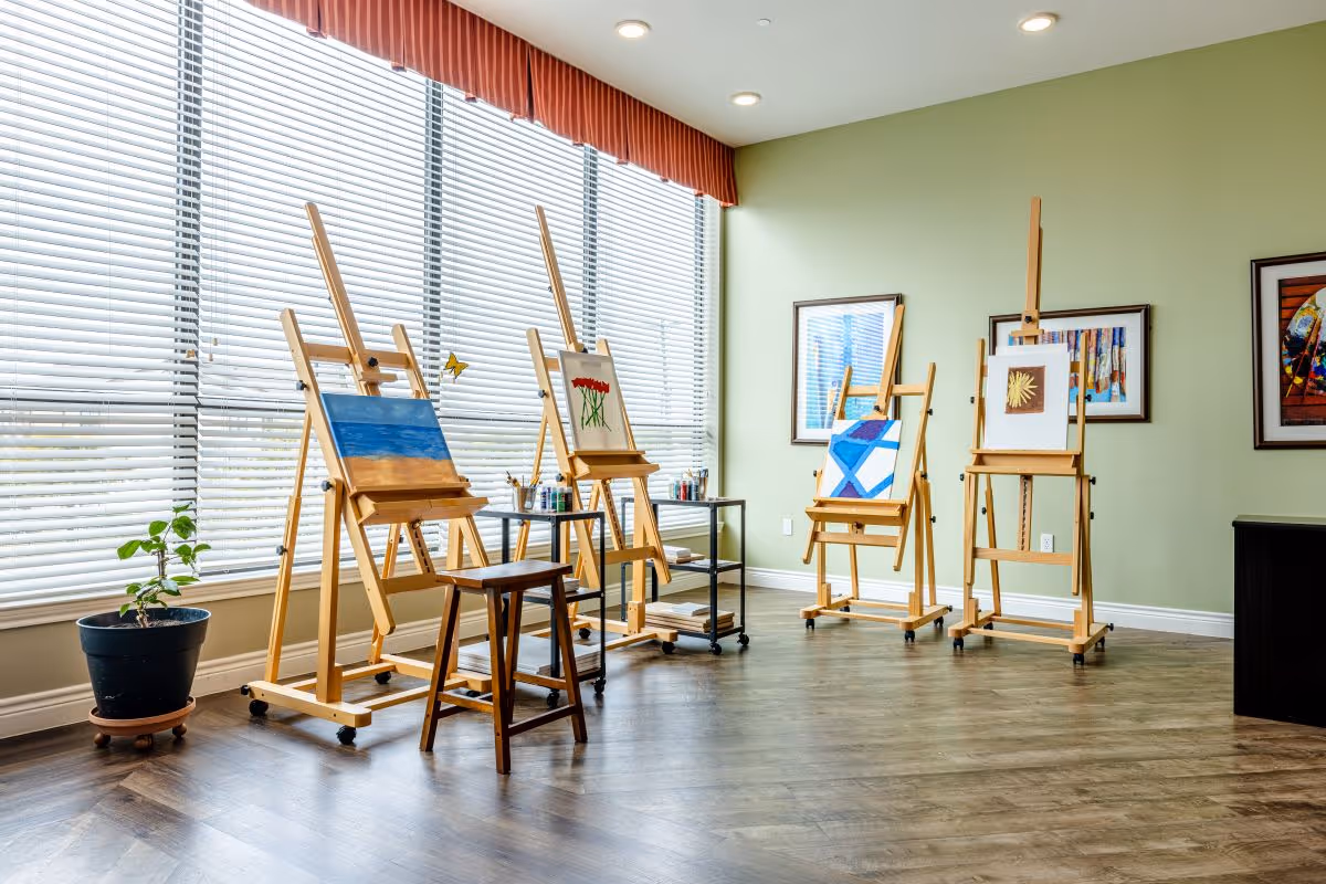 A bright art room with large windows covered by white blinds and a red valance. Four wooden easels display paintings, including a blue and orange abstract, red flowers, a blue geometric design, and a yellow flower. A wooden stool and a small black cart with art supplies are also present. The room has light green walls, framed artwork, and wood flooring.