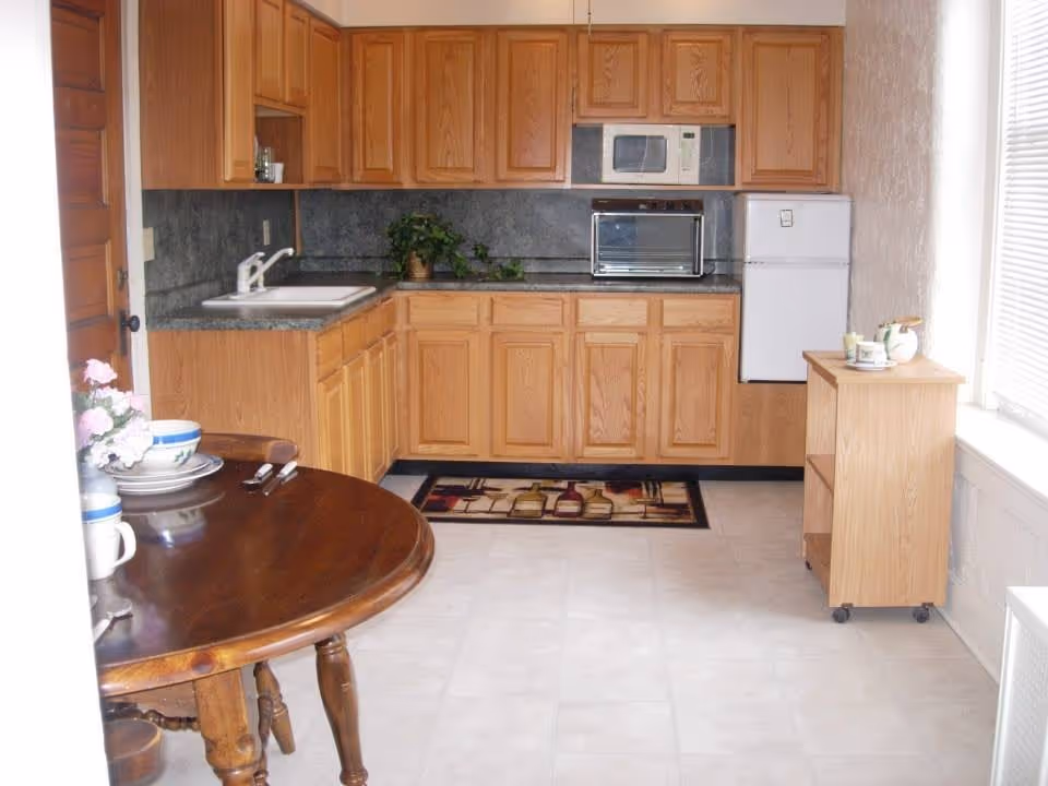 A small kitchen area with wooden cabinets, a white sink, a microwave, a toaster oven, and a compact white refrigerator. There is a wooden table with dishes and utensils on the left side and a small wooden cart with cups and a teapot on the right near a window with blinds. The floor is tiled and there is a decorative rug in front of the cabinets.