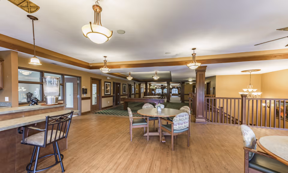 Open senior living common room with round dining tables and chairs, a service counter with stools, and a distant lounge area.