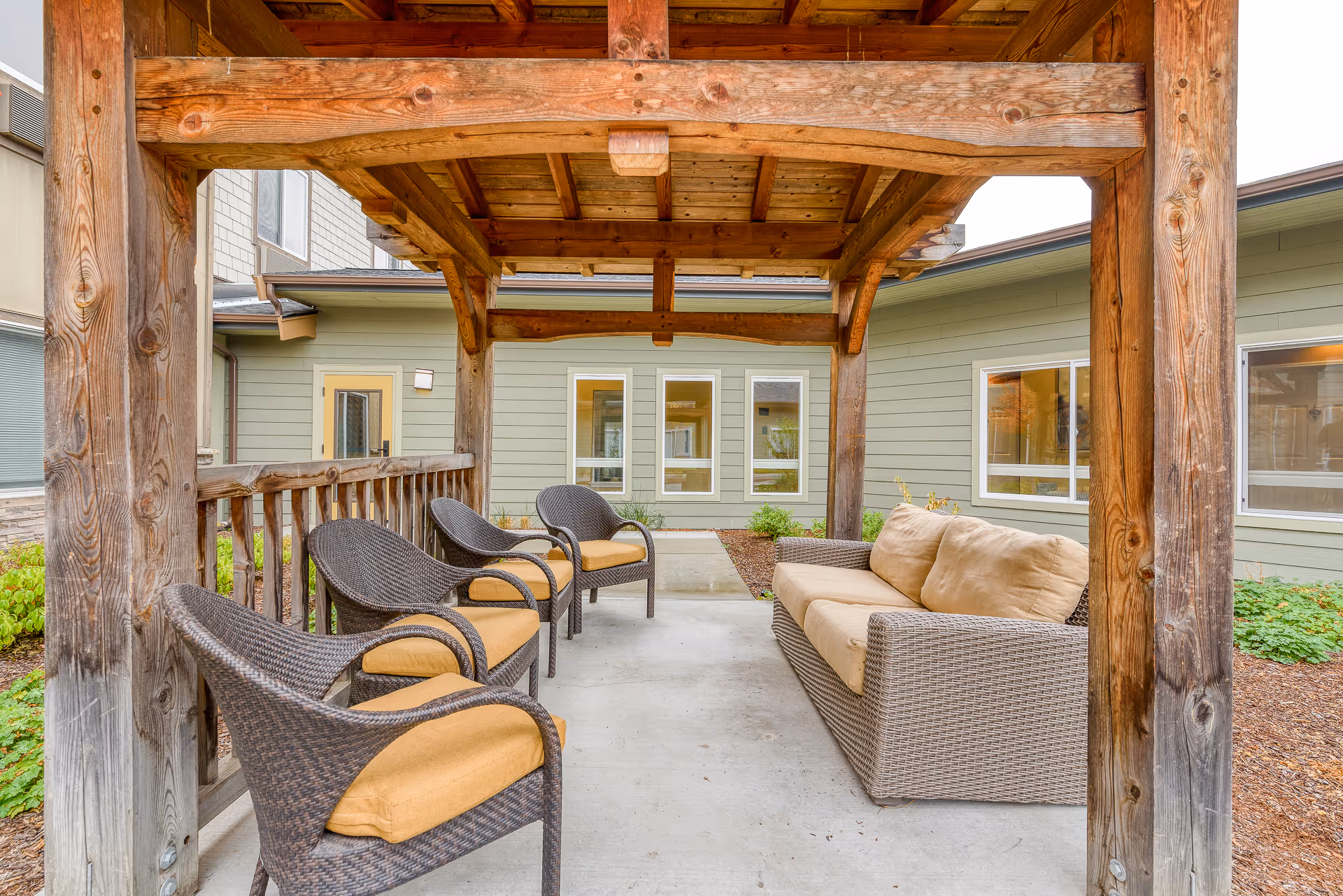 Outdoor covered seating area with a wooden pergola roof, featuring four wicker chairs with yellow cushions on the left and a wicker sofa with beige cushions on the right, surrounded by a garden and green building walls with windows.