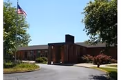 Brick single-story senior care facility with curved driveway, flag, trees, and landscaping.