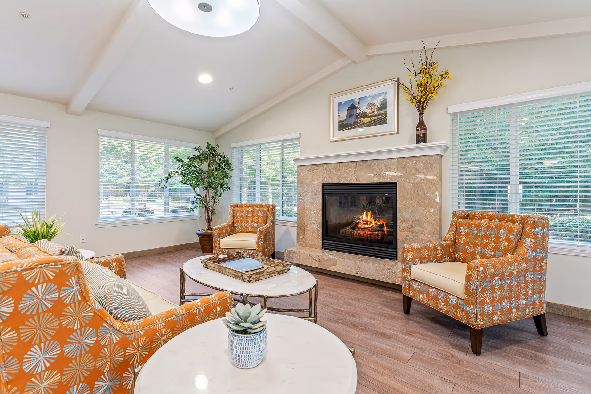 Bright communal living room with a lit fireplace, patterned orange armchairs, a sofa, coffee tables, and large windows.