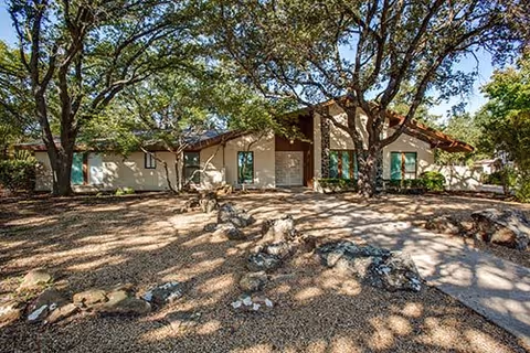 Single-story building with a beige exterior surrounded by large trees and a rocky, gravel-covered front yard under a clear blue sky.