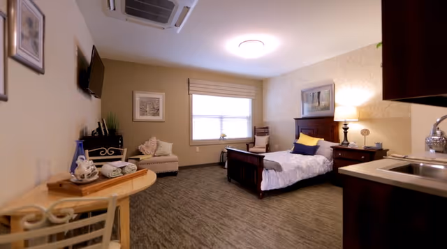 A cozy assisted living bedroom featuring a single bed with white bedding and pillows, a wooden nightstand with a lamp, a small armchair near the window, a wall-mounted TV, a small table with a tea set and towels, and a kitchenette area with a sink and cabinets.