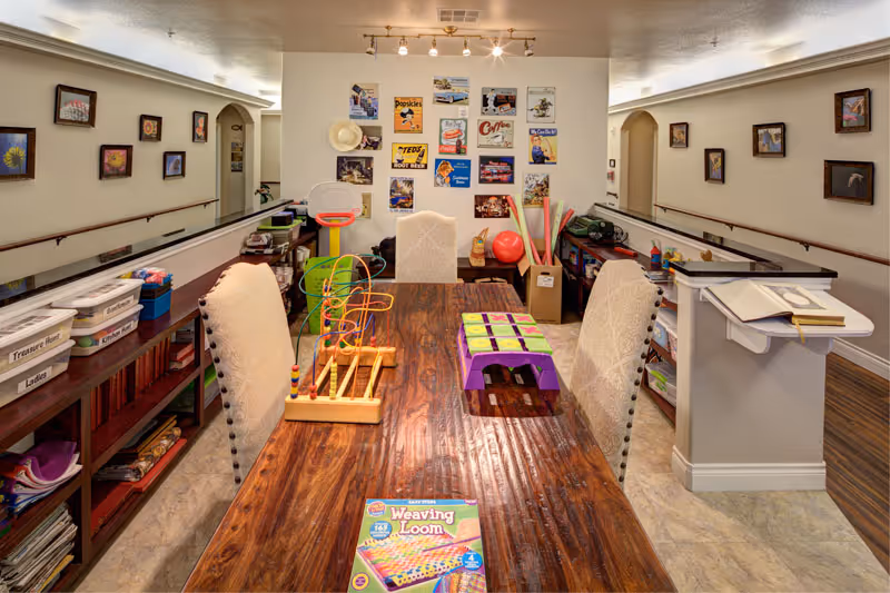 A well-lit activity room with a long wooden table surrounded by cushioned chairs. On the table are various games and craft supplies, including a weaving loom kit and a bead maze toy. The walls are decorated with framed pictures and colorful vintage-style posters. Shelves along the sides hold labeled bins and additional supplies. The room has a warm and inviting atmosphere, suitable for group activities.
