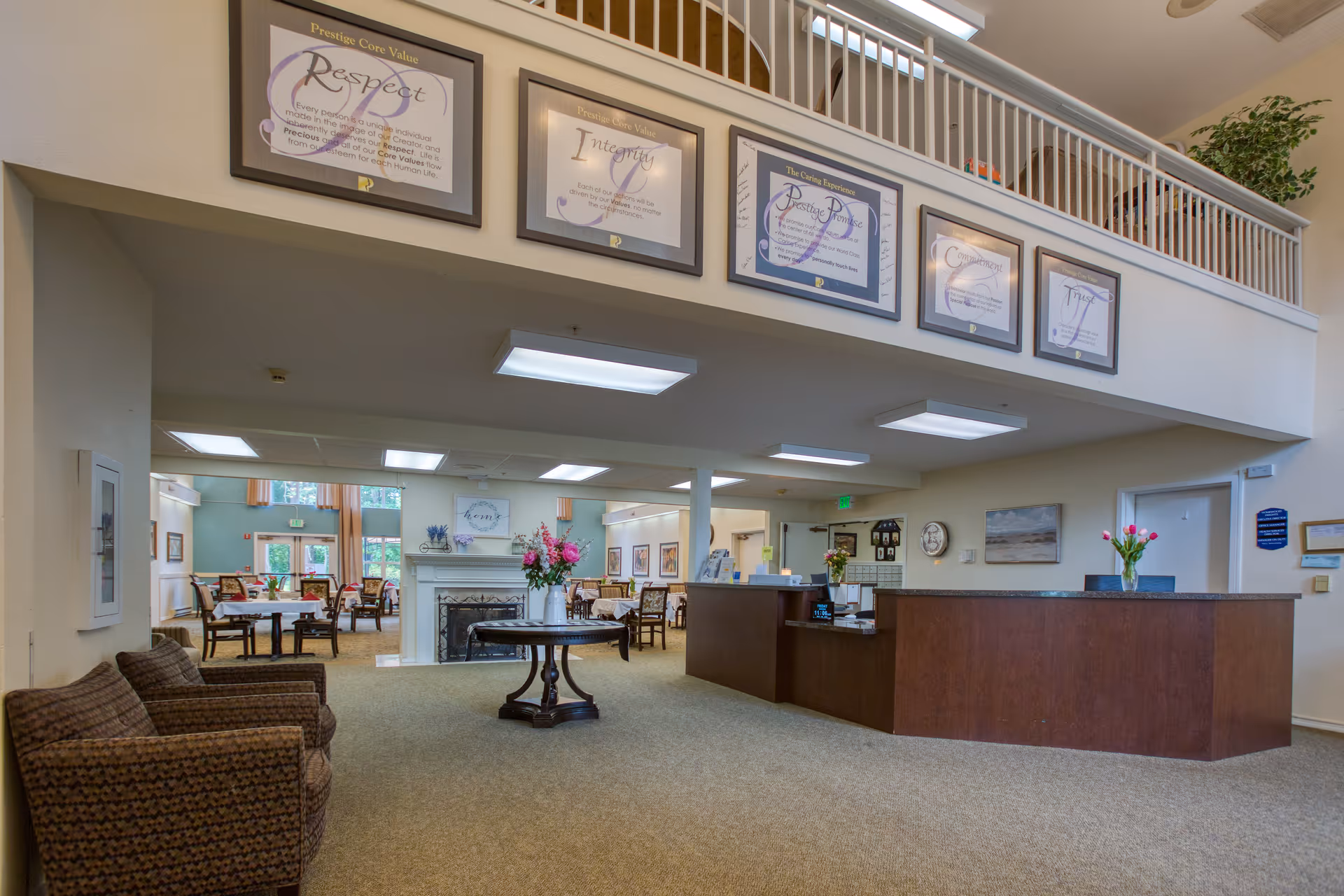 Bright interior lobby of an assisted living facility showing a reception desk, seating area, round table with flowers, and a dining area beyond.