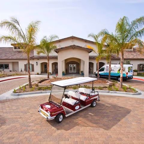 Entrance of a single-story senior living facility with a covered porte-cochère, palm trees, and a red electric shuttle parked in front.