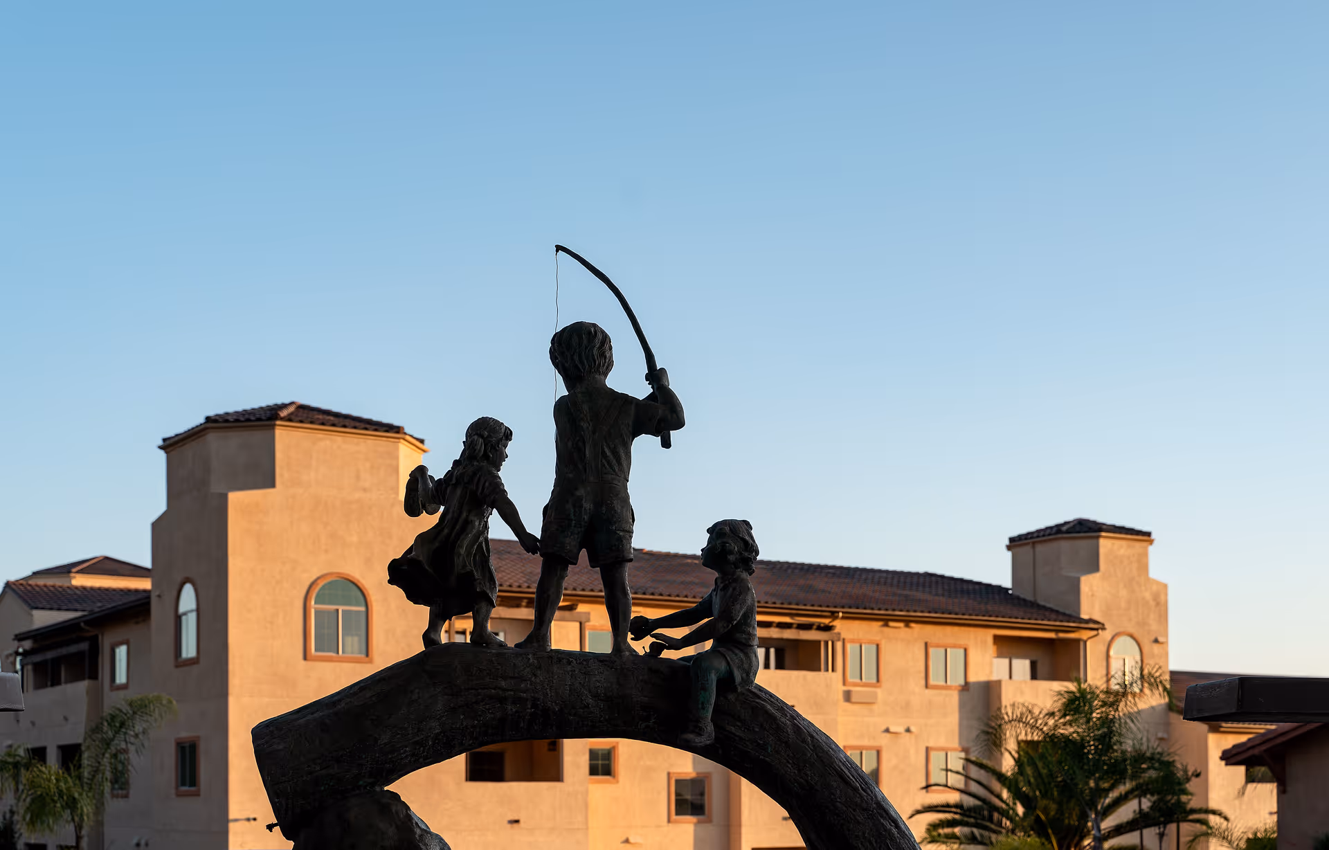 Bronze statue of three children playing on a curved log, one holding a fishing rod, set against the backdrop of a multi-story building with a tiled roof and palm trees under a clear blue sky.