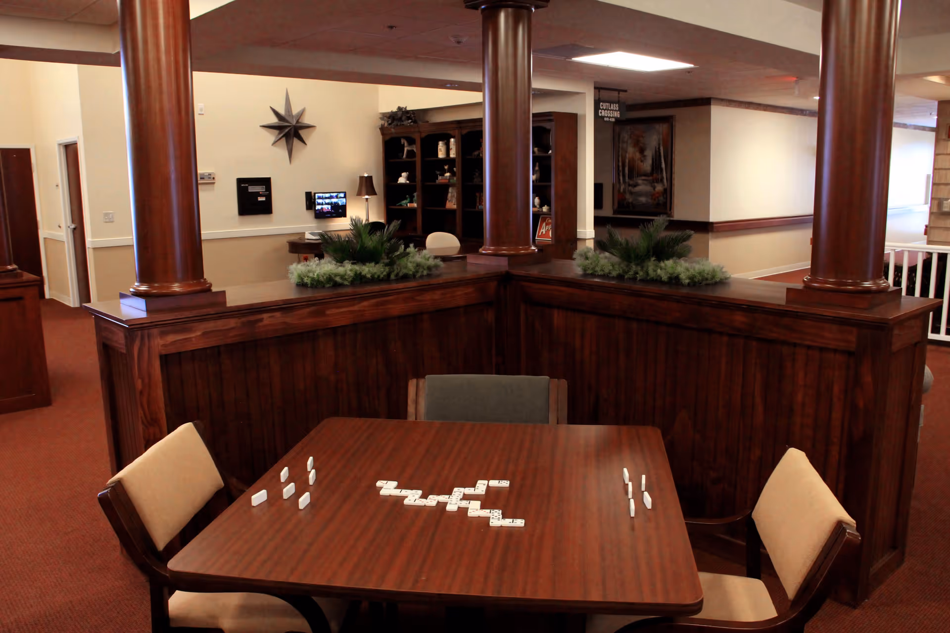 A cozy indoor common area with a wooden table and four chairs arranged around it. Domino tiles are set up on the table as if a game is in progress. The space features dark wood paneling and columns, with green plants on the divider behind the table. In the background, there is a bookshelf, a lamp, a wall-mounted screen, and a hallway with a sign reading 'Cutlass Crossing'.
