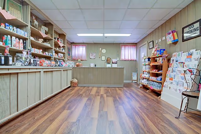 Interior view of a small shop or reception area with wooden flooring and paneled walls. Shelves on the left and right sides are stocked with various items including toiletries, snacks, and greeting cards. A counter is positioned at the back of the room beneath two small windows with pink curtains. A 'No Smoking' sign is visible on the right wall.