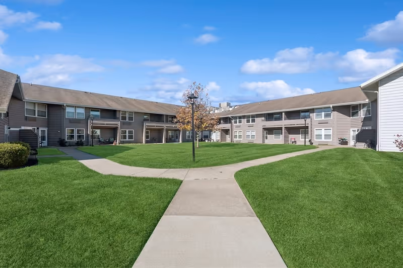 Outdoor courtyard area of a senior living facility with green grass, a concrete walkway, a few small trees, and two-story buildings with balconies and windows under a partly cloudy blue sky.