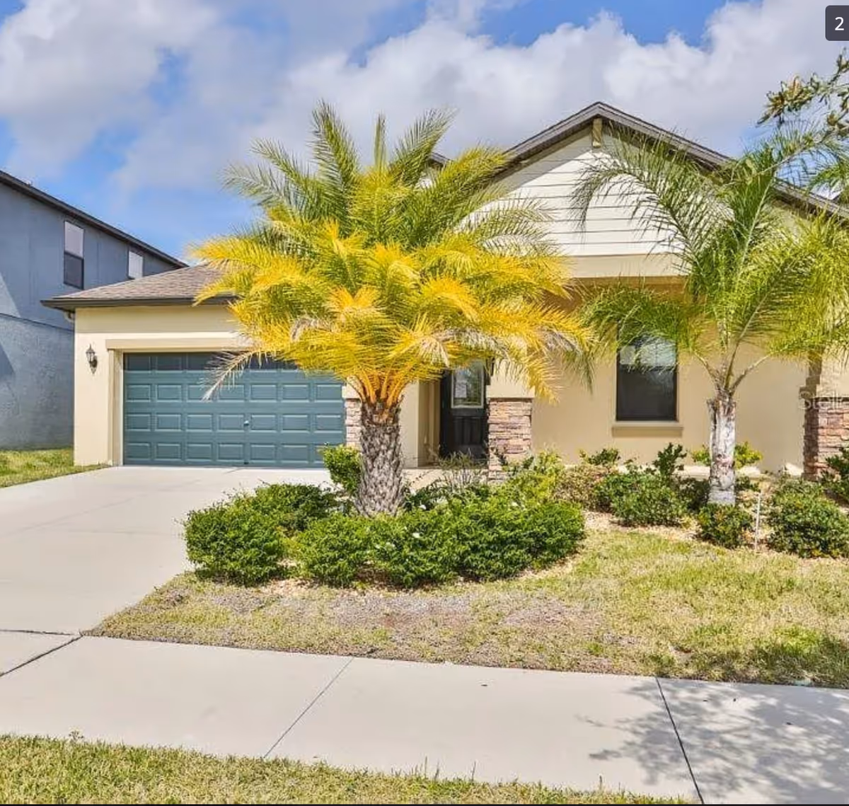 Front exterior of a single-story house with a two-car garage, palm trees, and landscaped yard.