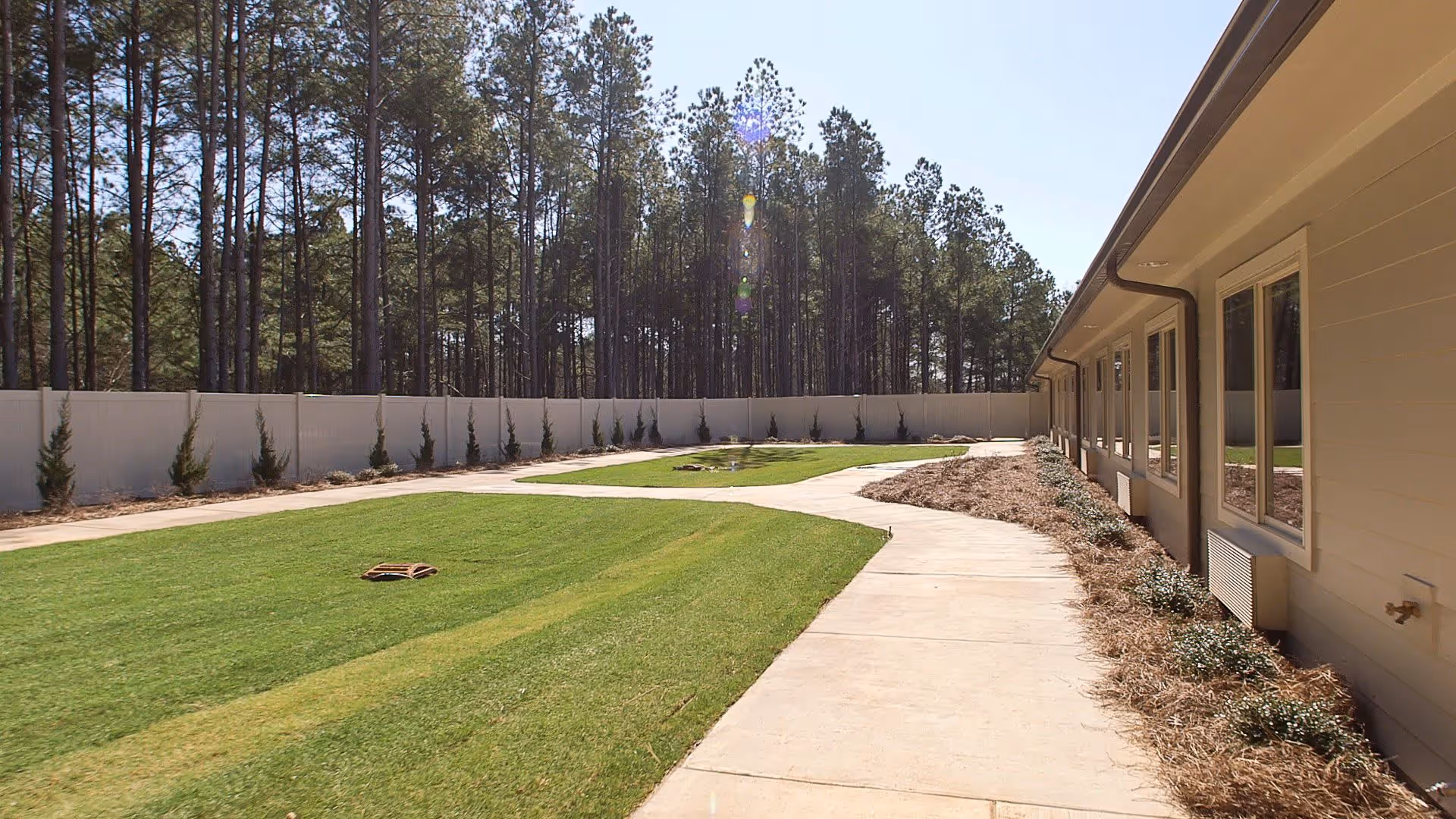 Outdoor view of a senior living facility showing a paved walkway curving alongside a building with multiple windows. There is a well-maintained grassy area bordered by a white fence and tall pine trees in the background under a clear sky.