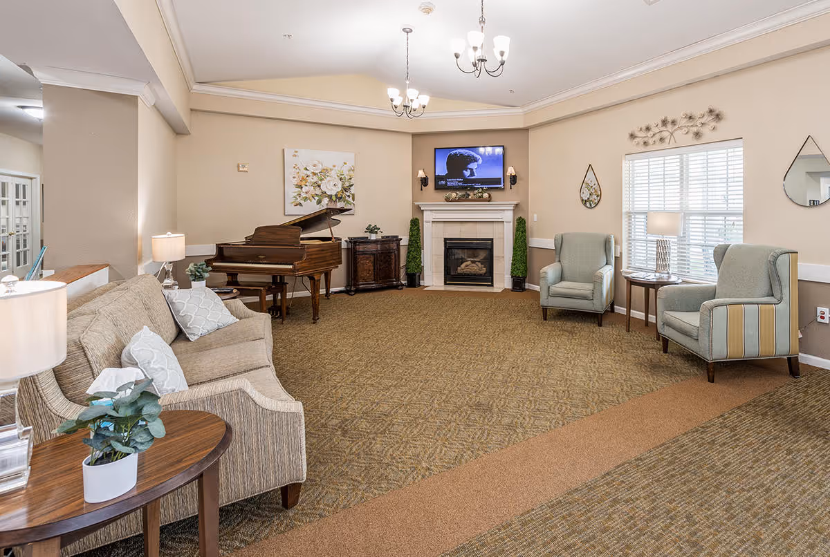 A cozy living room area in Digby Place featuring a beige sofa with patterned pillows, two light blue armchairs, a wooden grand piano, a fireplace with a TV mounted above it, and decorative wall art. The room is well-lit with ceiling lights and table lamps, and has a carpeted floor.