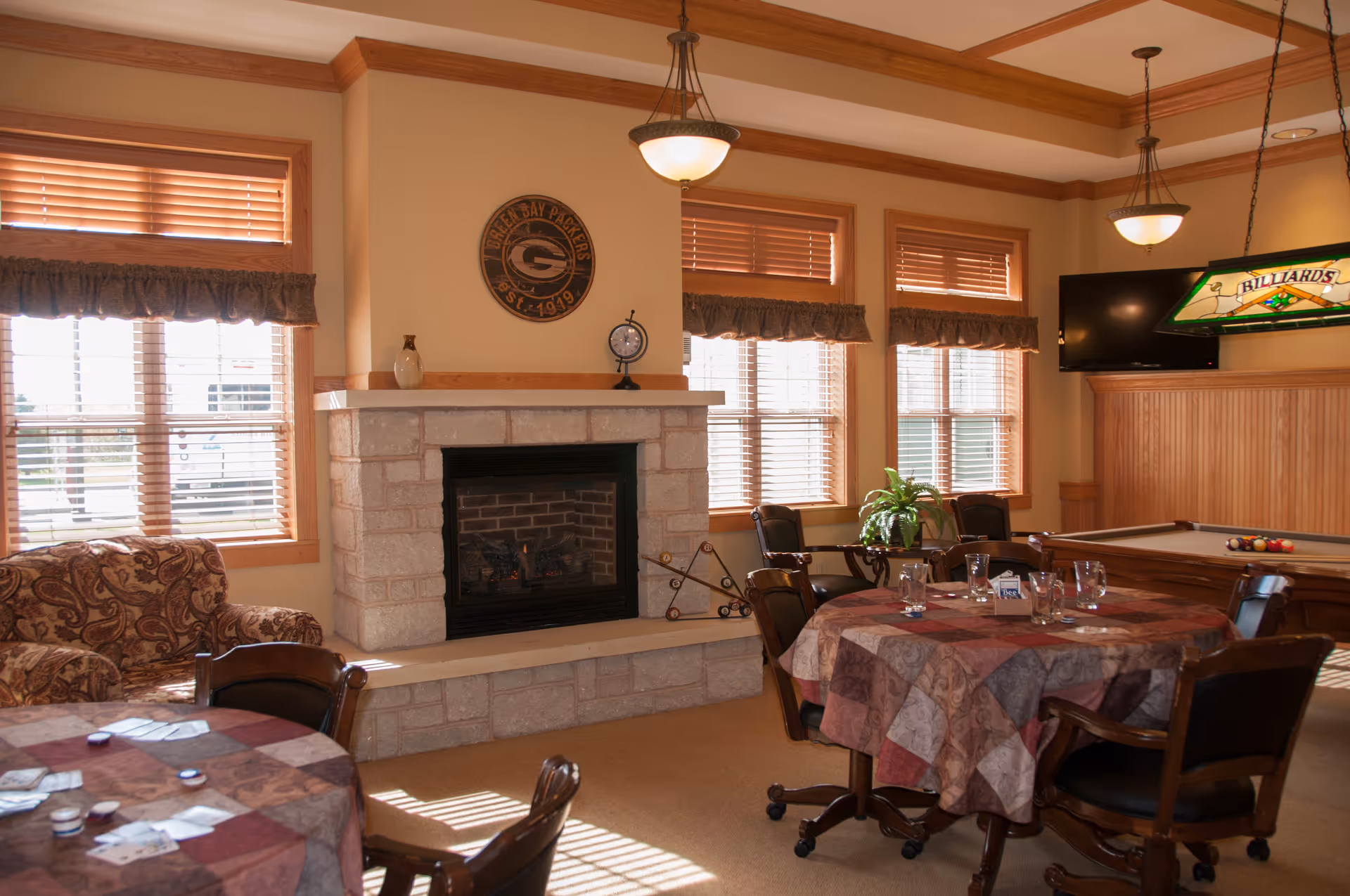 A cozy common area with a stone fireplace, multiple windows with wooden blinds, a patterned armchair, round tables covered with patchwork tablecloths, chairs on wheels, a billiards table with a stained glass light fixture above it, and a wall-mounted TV.
