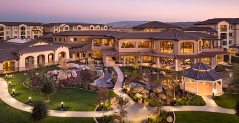 Twilight exterior view of a multi-story senior living complex overlooking a landscaped courtyard with a pond, gazebo, and winding walkways.