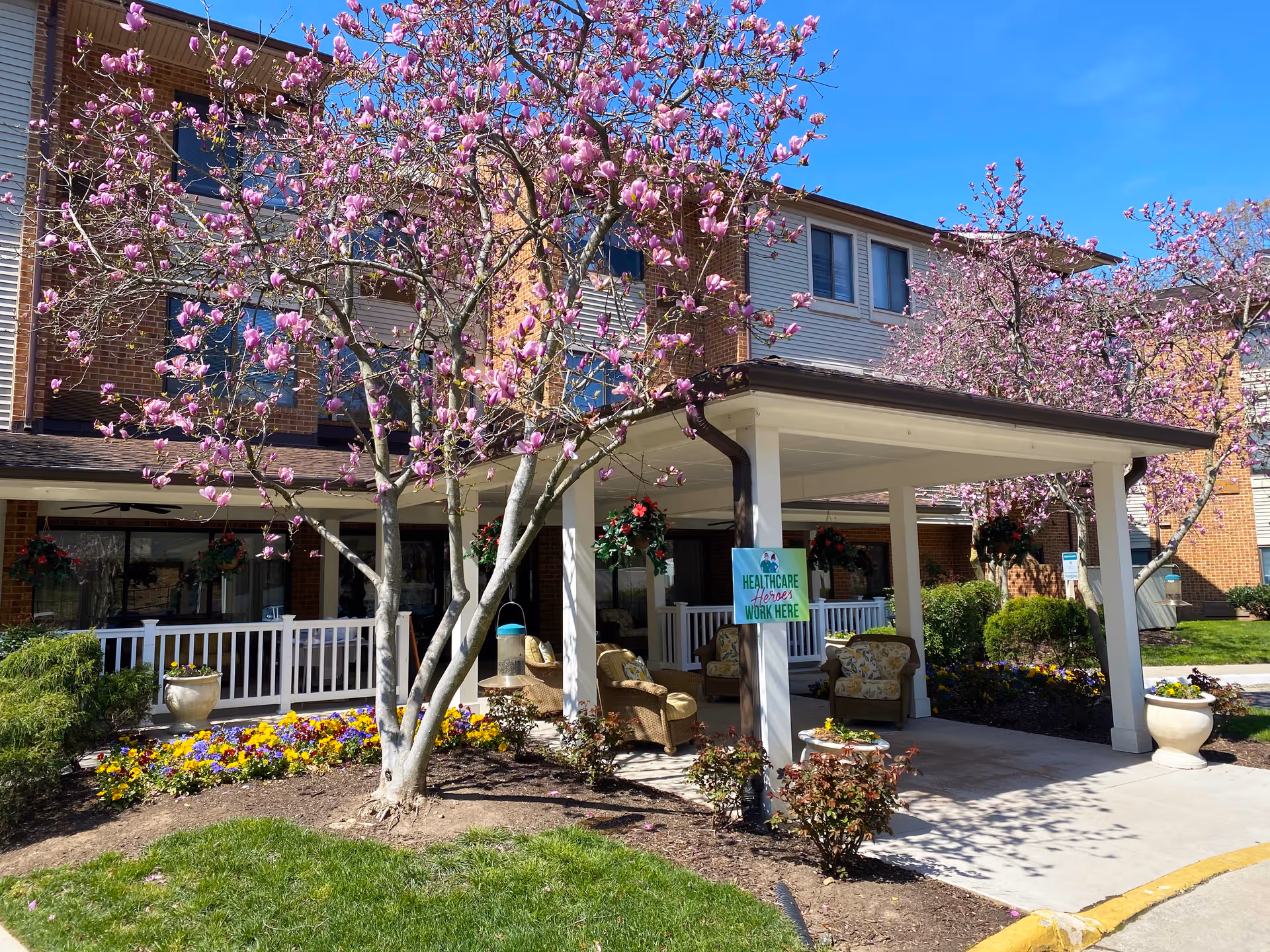 Exterior view of Potomac Place Assisted Living and Memory Care building with blooming pink magnolia trees and colorful flower beds. There is a covered entrance with outdoor seating including cushioned chairs and hanging flower baskets. A sign on one of the pillars reads 'Healthcare Heroes Work Here'.