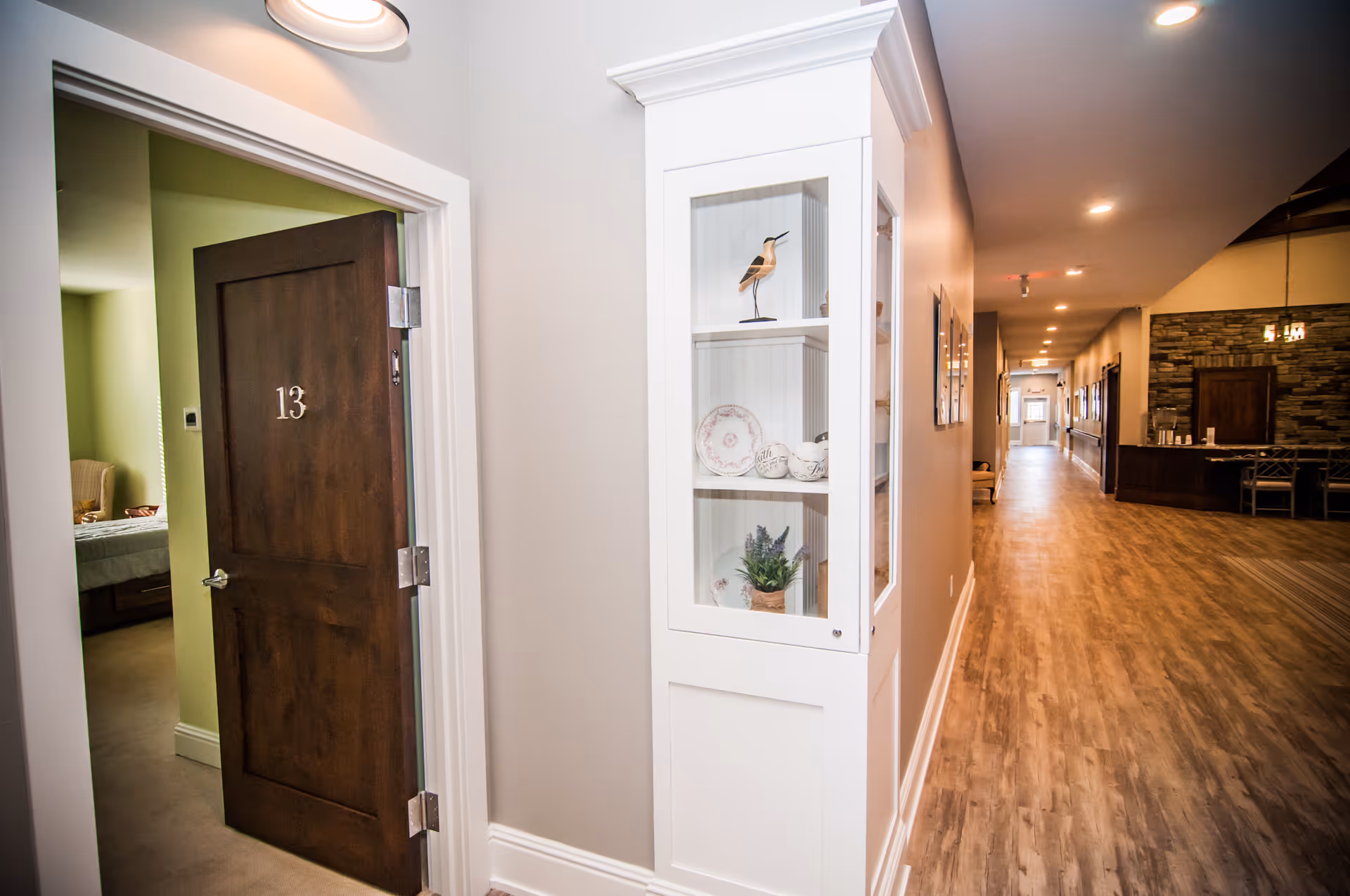 A hallway in a senior living facility with wooden flooring and recessed lighting. On the left, there is an open door labeled number 13 leading to a room with a bed and chair. A white built-in display cabinet with decorative items is mounted on the wall. At the end of the hallway, there is a seating area with a stone accent wall and a table with chairs.