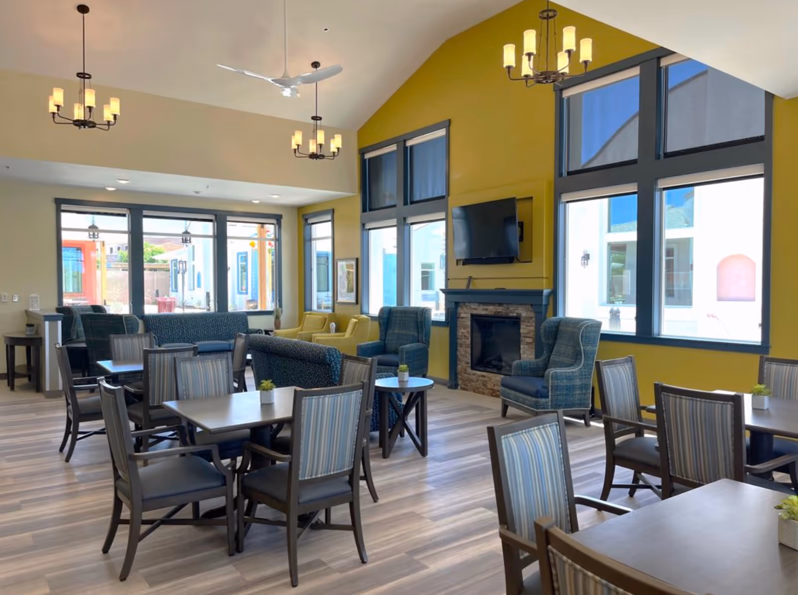A bright and spacious common area with multiple tables and chairs arranged for seating. The room features large windows allowing natural light to fill the space, a yellow accent wall with a mounted flat-screen TV above a stone fireplace, and comfortable armchairs and sofas. Ceiling fans and chandeliers hang from the high ceiling, and small potted plants decorate the tables.