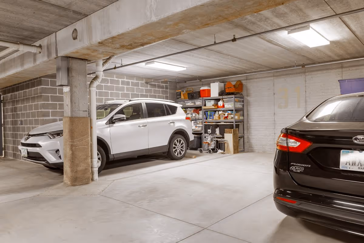 Indoor parking garage with two parked cars, one white SUV and one black sedan. There is a concrete pillar wrapped with padding and shelves with various storage items against the wall.