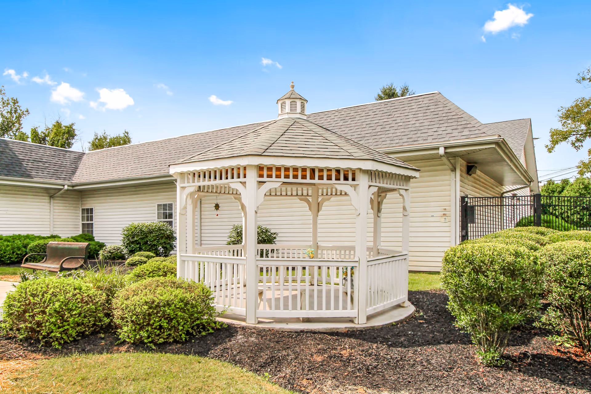 White wooden gazebo in a landscaped courtyard in front of a light-colored building under a blue sky.