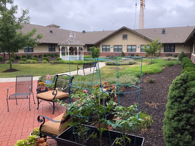 Outdoor garden area at Morningside Place featuring patio chairs with cushions, a small garden with plants supported by metal cages, a brick-paved walkway, a white gazebo in the background, and a building with multiple windows under a cloudy sky.