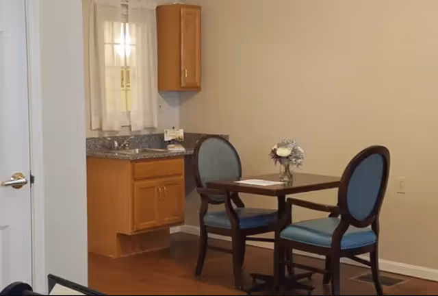 A small dining area with a wooden table and two blue cushioned chairs. In the background, there is a kitchenette with wooden cabinets, a granite countertop, a small sink, and a window with sheer white curtains. The walls are beige and the floor is wooden.