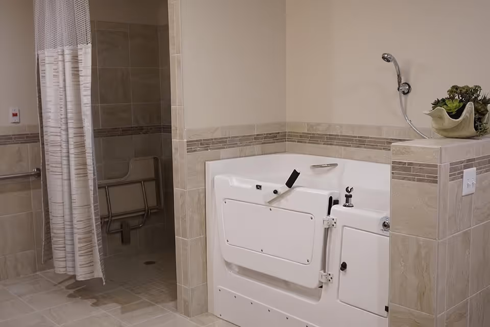 A bathroom with beige tiled walls and floor featuring a walk-in shower with a safety grab bar and a white curtain. Next to the shower is a white walk-in bathtub with a door and a handheld showerhead. A small plant in a decorative pot is placed on the tiled ledge beside the bathtub.