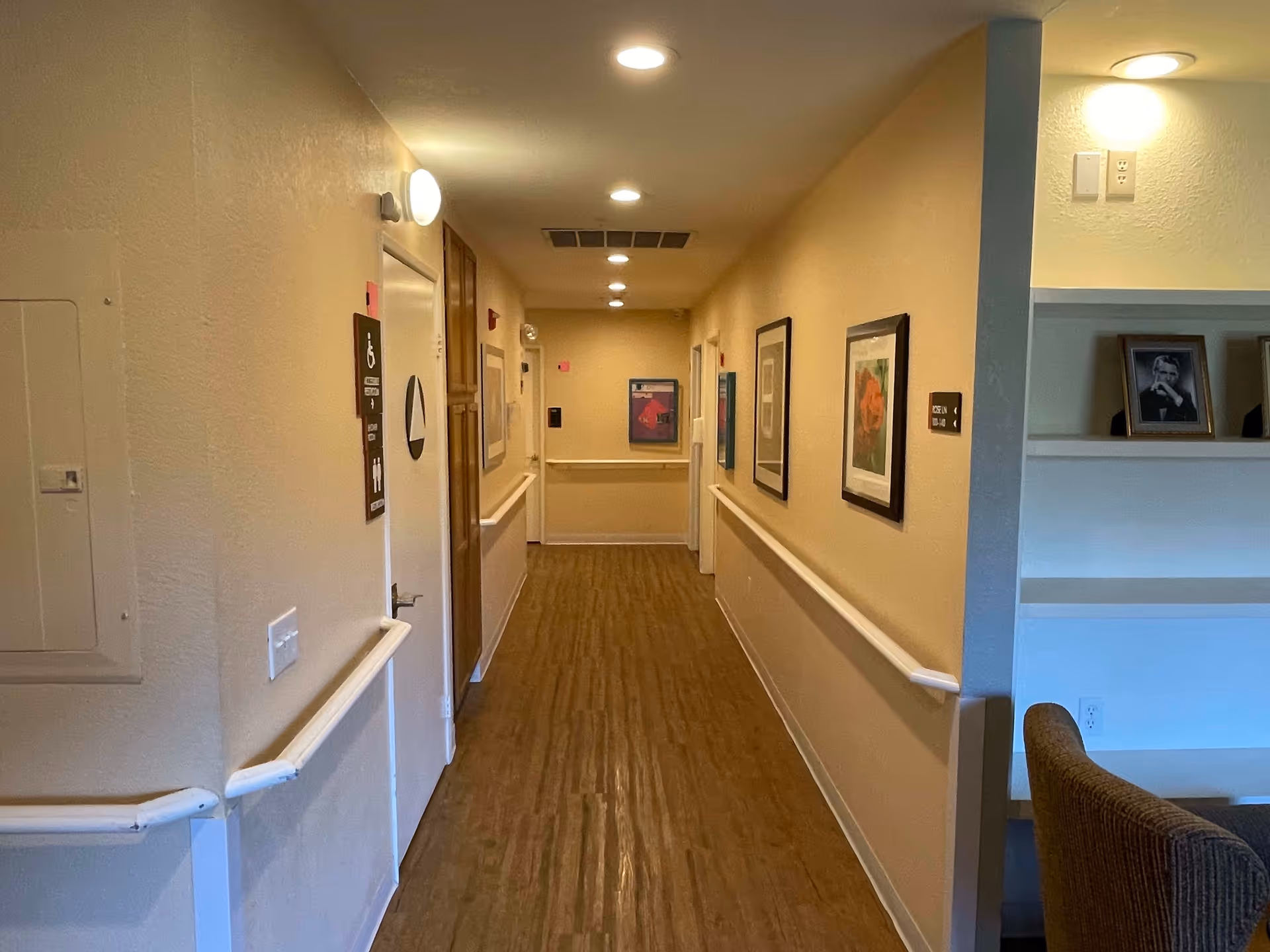 A well-lit hallway in a senior living facility with wooden flooring, handrails on both sides, framed artwork on the walls, and a door marked with a handicap accessible restroom sign on the left side.