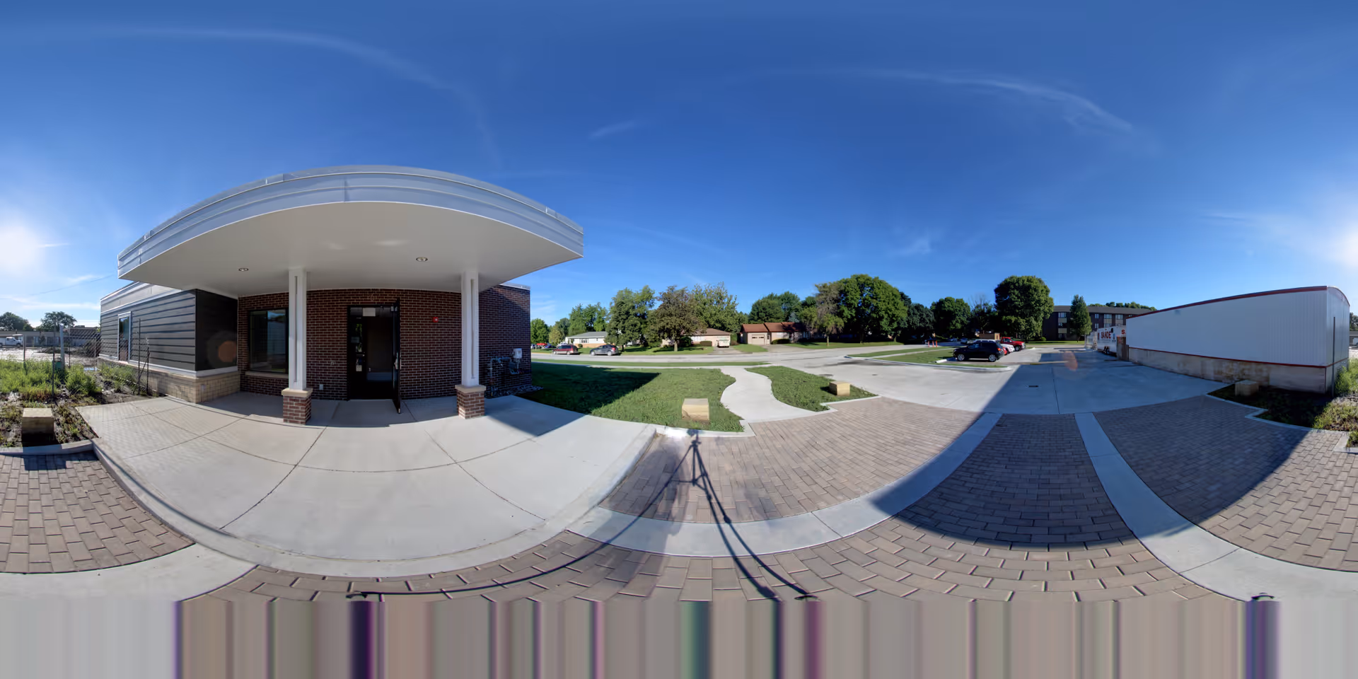 Wide panoramic view of the exterior entrance of a building with a covered porch supported by two white columns. The building has brick and siding walls. There is a paved walkway and parking area with some cars visible. Trees and a clear blue sky are in the background.