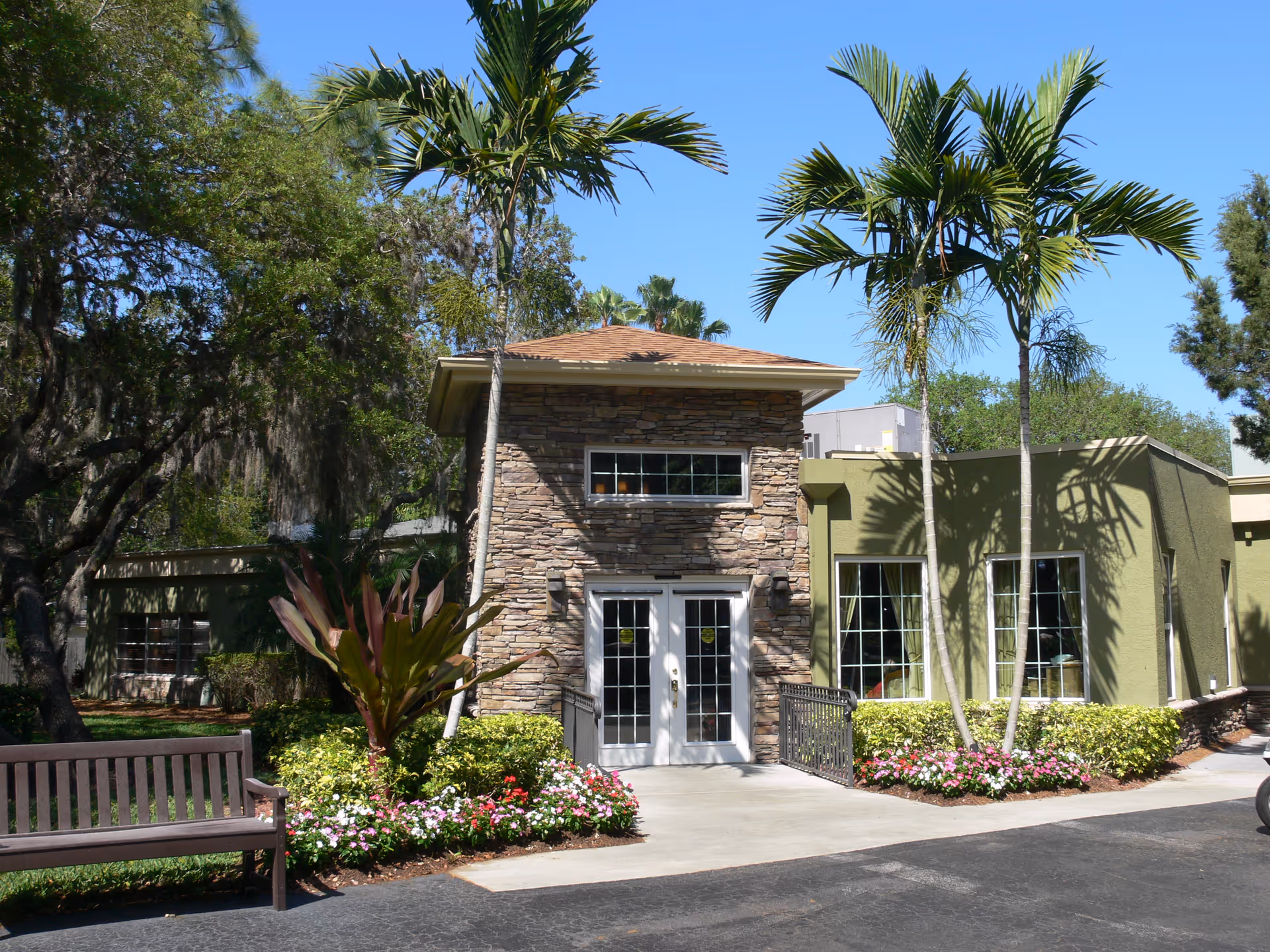 Entrance of a senior living building with a stone facade, glass double doors, palm trees, bench and flower beds under a clear blue sky.