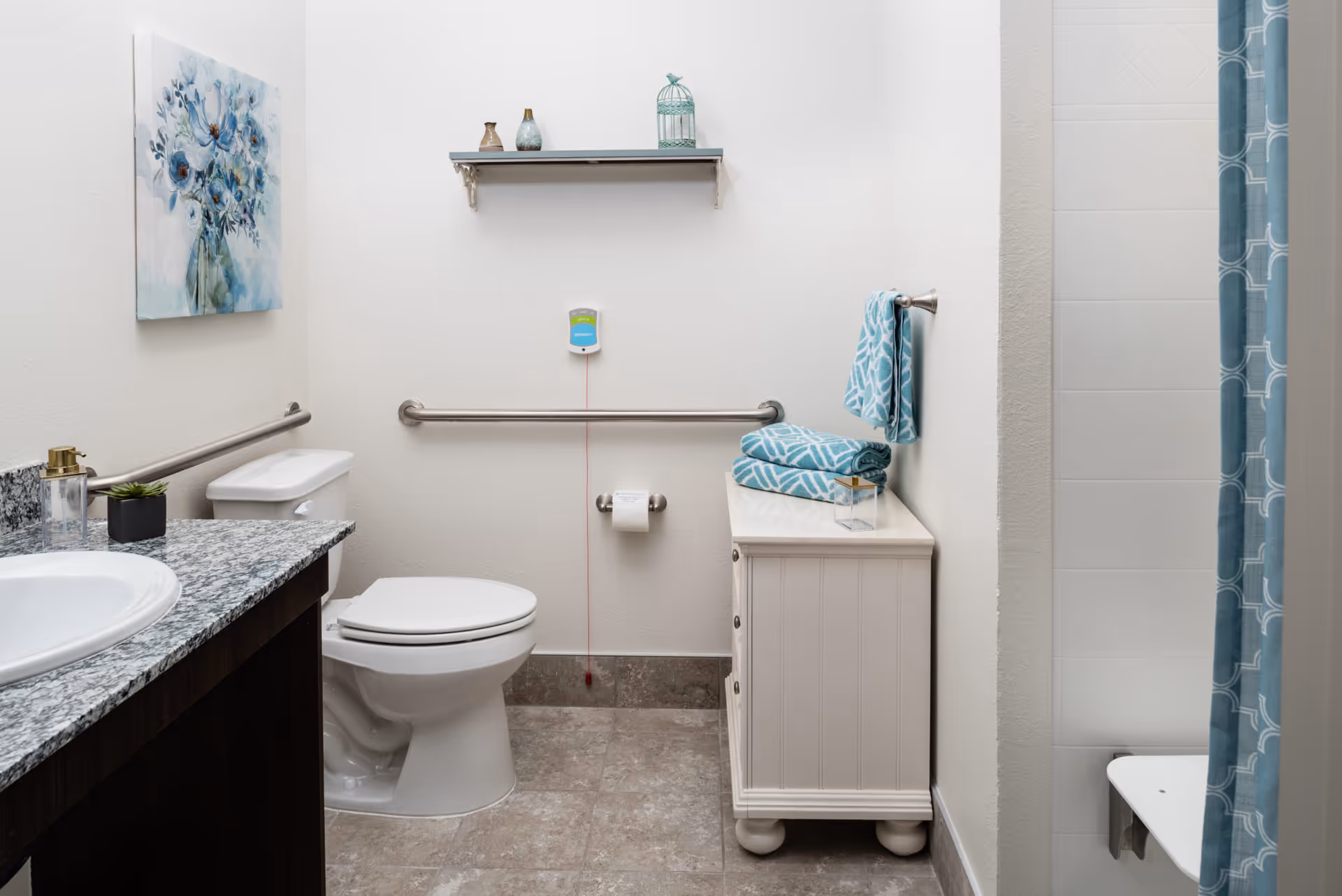 A clean and well-organized bathroom with a toilet, a sink with granite countertop, a white cabinet with folded blue patterned towels, a shower with a blue patterned curtain, a wall-mounted shelf with decorative items, and safety grab bars on the walls.